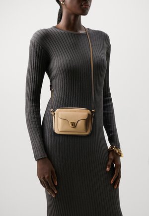 Beige lædertaske med tværrem, buet klap, guldfarvet hardware og en tekstureret overflade, båret over en ribstrikket grå kjole.