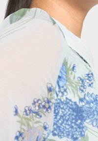 Blouse légère et transparente avec un motif floral bleu et vert, présentant une texture subtile. Le design intègre des fleurs et des feuilles vibrantes.