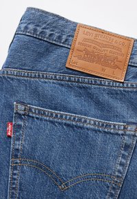 Jean Levi's en denim bleu avec poche arrière visible, étiquette rouge Levi's, coutures orange et étiquette de marque en cuir marron à la taille.