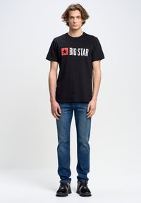 Juodas medvilninis marškinėlis su balta ir raudona „BIG STAR“ logotipu. Sujuostas su mėlynomis džinsinėmis kelnėmis ir juodais sportbačiais. Standartinis pjūvis.