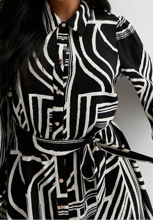 Nainen, joka käyttää mustavalkoista, geometrisella kuvioilla varustettua nappisuljettavaa mekkoa, jossa on solmittava vyö vyötäröllä ja pitkät hihat.