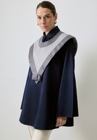 Cappa bleumarin cu guler lat gri și detaliu cu fundă în față. Dispune de mâneci lungi, cu o textură netedă și un design minimalist.