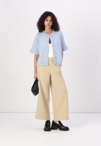 Cardigan bleu clair à manches courtes avec décolleté rond et fermeture à boutons, associé à un pantalon large beige et des mocassins noirs, tenant un sac noir.