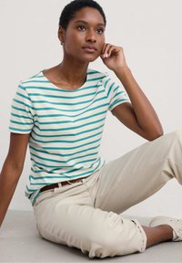 Camiseta de rayas en teal claro y crema, de manga corta, con cuello redondo, fabricada en suave algodón. Lleva puestos pantalones beige y zapatos claros.