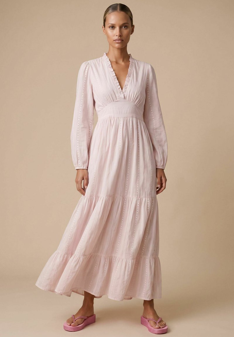 Femme portant une longue robe rose clair avec un décolleté en V profond et des manches longues, associée à des sandales compensées roses, debout sur un fond beige.