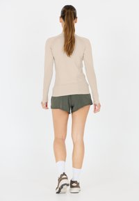Beige langærmet top i struktureret stof, parret med grønne shorts. Model iført lyse sneakers og hvide sokker, set bagfra.