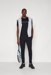 T-shirt noir avec texte blanc, hoodie sans manches gris, jog pants noirs et baskets noires. Tenant une raquette de tennis avec des accents bleus.