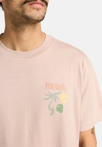 T-shirt en coton rose clair avec logo RVCA brodé et un design comportant des feuilles de palmier, un soleil et un visage en accents verts et jaunes.