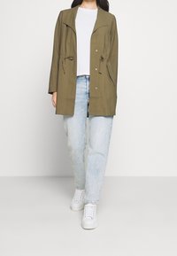 Olive green long-sleeve jacket with a collar, drawstring waist, snap buttons, and two side pockets, worn over a white shirt and light jeans.