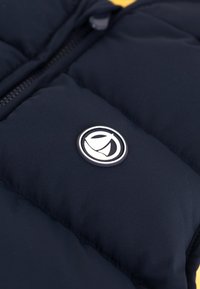 Tamnoplavi pufasti prsluk s srebrnim logotipom, glatkom teksturom tkanine i prednjim zatvaračem.