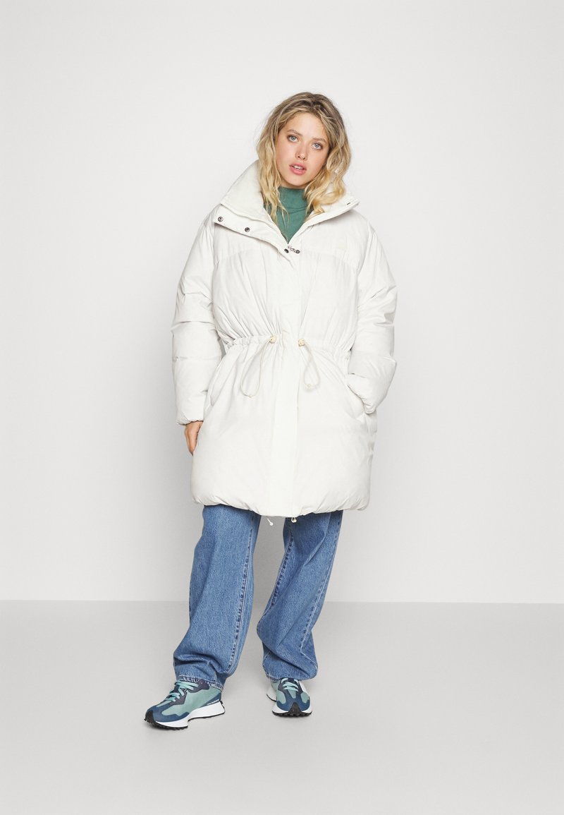 Abrigo de plumas blanco con cintura ajustable, cuello alto y detalles de bolsillo con cremallera. Llevado sobre un suéter verde y combinado con jeans azules y zapatillas.