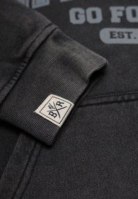 Tummanharmaa collegekangas, jossa on teksturoitu pinta, mukana valko-mustalla logopatchilla hihassa ja painettuna tekstimuotoiluna.