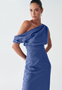 Žena se sebevědomě prezentuje ve stylových modrých šatech s jedním ramenem, které zdůrazňují její eleganci na světlém pozadí.