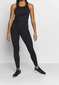 Musta urheilu-toppi, jossa on kierretty solmu-yksityiskohta, yhdistettynä tiukkoihin mustiin leggareihin. Mustat urheilukengät täydentävät asua.