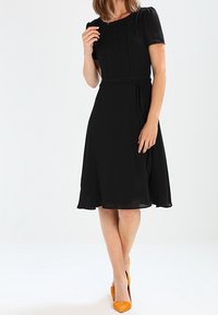 Černé šaty po kolena s krátkými rukávy, detail nařaseného korzetu a vázaným pasem. Spárovány s oranžovými lodičkami s špičatou špičkou. Hladká textura.