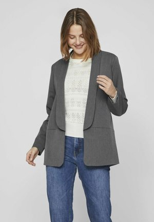 Veste cintrée grise avec un col cranté, devant ouvert et deux poches avant, portée par-dessus un léger pull en tricot et un jean bleu.