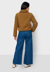 Hnědá, texturovaná, umělá kožichová bunda s límcem, zkombinovaná s modrými širokými džíny s vysokým pasem a světlou obuví. Zobrazený pohled zezadu.