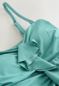 Türkizkék ujjatlan ruha fodros díszítéssel, sima textúrával és állítható pántokkal. Az anyag lágyan omlik, elegáns kialakítást mutatva.