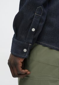 Rękaw ciemnej kurtki dżinsowej z srebrnymi zatrzaskami, kontrastującym przeszyciem i dopasowanym krojem, w połączeniu z zielonym materiałem pod spodem.