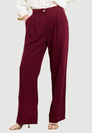 Kobieta nosząca bordowe, szerokie spodnie z wysokim stanem i bocznymi kieszeniami, zestawione z białą bluzką i złotymi odkrytymi szpilkami.