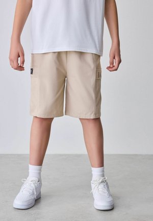 Beige Shorts mit zwei Seitentaschen und einem Etikett auf der linken Seite, getragen mit einem weißen T-Shirt und weißen Sneakern mit gerippten Socken.