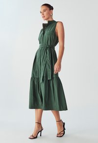 Roheline, varrukateta, kraega kleit, millel on kitsendatud vöökoht ja kihiline seelik. Omab nööpe ja seotud vööd; kantud mustade rihmadega kontsadega.