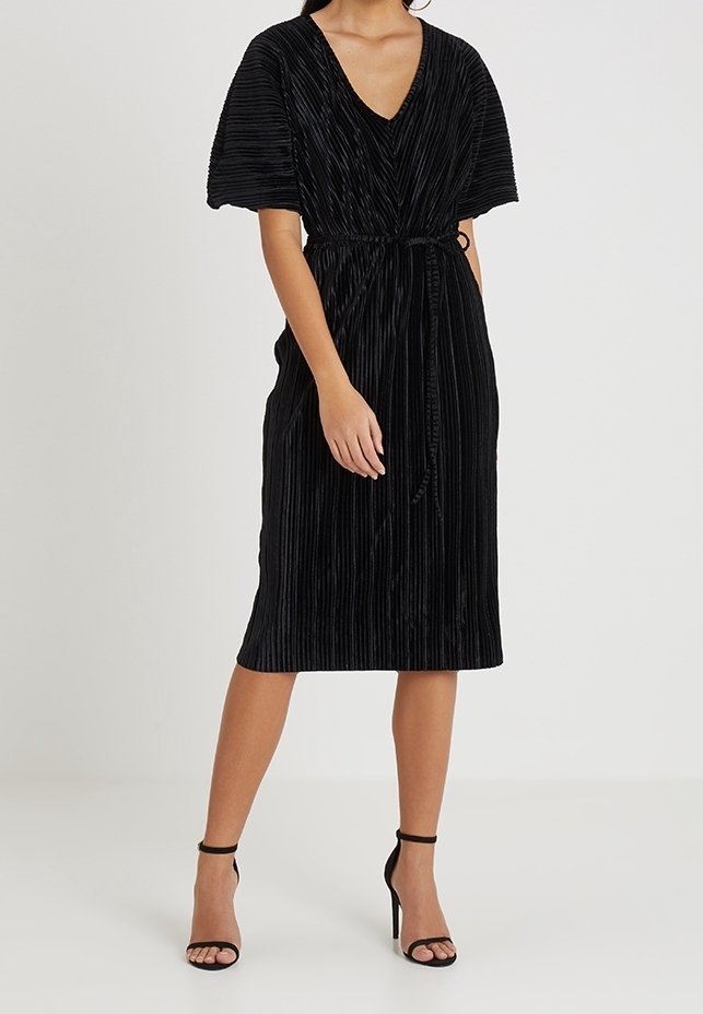 Femme portant une robe portefeuille plissée noire longueur genou avec ceinture à la taille et manches volantées, associée à des sandales à talons hauts noires.