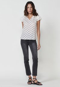 Blusa bianca a pois con maniche a volant, abbinata a jeans neri e sandali con cinturino, che mostra una silhouette casual e aderente.