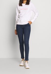 Felpa bianca con logo rosa "Lee Jeans", abbinata a jeans skinny blu scuro e sneakers bianche, in piedi davanti a uno sfondo neutro.