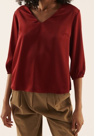 Vrouw draagt een losse rode V-hals blouse met driekwart mouwen en beige geplooide broek, staand tegen een lichte achtergrond.