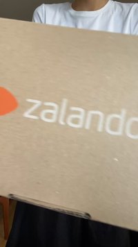 Bruine kartonnen verpakking met witte "zalando" tekst en een oranje logo bovenop, met een eenvoudige rechthoekige vorm en een gestructureerd oppervlak.