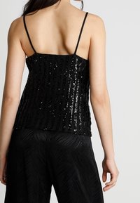 Svart paljett-cami med tunna axelband, med vertikala randmönster. Tyget har en glansig textur, som kombineras med vida svarta byxor.
