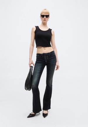 Femme portant un crop top noir sans manches, un jean évasé foncé, des talons pointus noirs, des lunettes de soleil, tenant un sac en cuir noir devant un fond blanc uni.