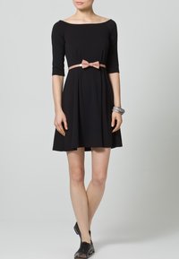 Czarna sukienka z rękawami o długości trzech czwartek, dopasowanym korpusie, rozkloszowanym dołem i jasnoróżową kokardą w talii. Gładki materiał o minimalnej teksturze.