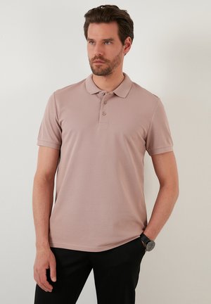 Svetlo mauve polo majica z zavihanim ovratnikom, trojnim gumbom in kratkimi rokavi. Teksturirana tkanina. Nosijo jo z temnimi hlačami in uro.