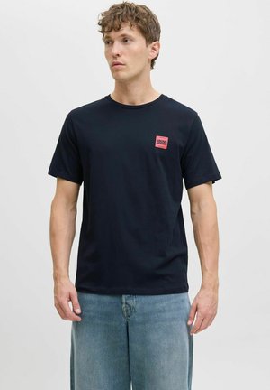 Sort bomulds t-shirt med et lille rødt firkantet logo på venstre bryst. Korte ærmer og rund halsdesign. Casual pasform.