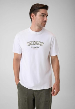 Mand med brunt hår iført en hvid Guess Country Club t-shirt og olivengrønne bukser, der kigger til højre med hænderne nær lommerne.