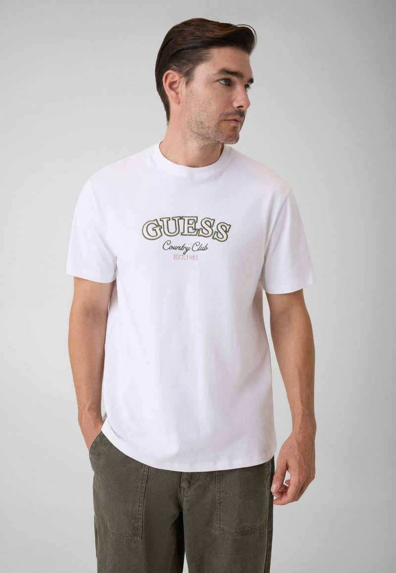 Άνδρας με καστανά μαλλιά που φοράει λευκό T-shirt Guess Country Club και χακί παντελόνια, κοιτάζοντας προς τα δεξιά με τα χέρια κοντά στις τσέπες.