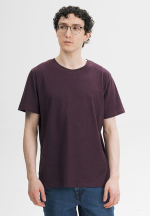 Jonge man met krullend donker haar en een bril, die een effen donkerpaars T-shirt en blauwe spijkerbroek draagt, kijkt iets opzij.