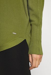 Pull en tricot vert olive, avec un design texturé, des poignets et un ourlet côtelés, un détail fente sur le côté et une étiquette de marque près de l'ourlet.