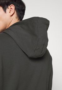 Sudadera de color gris oscuro con una textura suave, capucha con cordón y líneas simples y limpias. El tejido parece transpirable y casual.