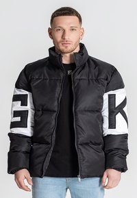 Chaqueta de plumas en blanco y negro con un alto cuello, cierre de cremallera y grandes acentos de logo en las mangas. Presenta una textura acolchada y una silueta inflada.