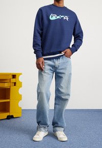 Mörkblå sweatshirt med turkos och rosa grafiskt logotyp, kombinerad med ljusblå jeans och vita sneakers. Rum med blå matta och gul förvaring.