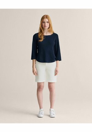 Kvinde med rødt hår iført en marineblå tekstureret langærmet top, hvide knælange shorts og hvide sneakers, stående mod en beige baggrund.