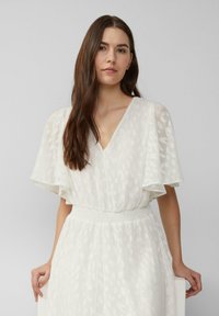 Giovane donna con lunghi capelli castani vestita con un abito bianco strutturato e maniche a drappo, in piedi contro uno sfondo grigio chiaro uniforme.