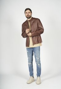 Brun läderjacka med ståkrage, med diagonal sömnad och två fickor med dragkedja. Bärs över en beige skjorta med trasiga blå jeans.