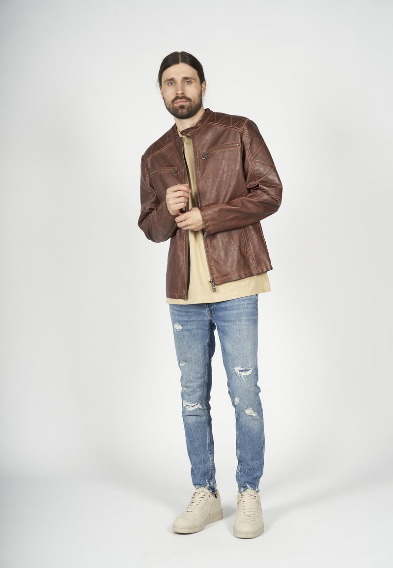 Veste en cuir brun avec un col montant, présentant des coutures diagonales et deux poches zippées. Portée par-dessus une chemise beige avec un jean bleu déchiré.