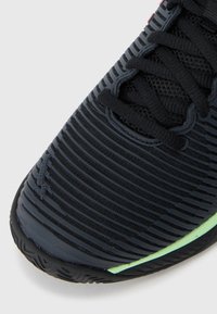 Black sports shoe with textured upper, featuring thin horizontal lines and a green accent on the sole. Laces are black and woven.