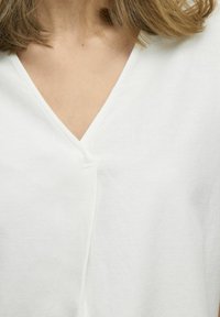 Weißes V-Ausschnitt-Oberteil mit strukturiertem Finish. Verfügt über eine subtile Nahtdetails in der Mitte und eine lockere Passform, die im Bereich des Ausschnitts sichtbar ist.