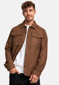 Braune Wildlederjacke mit Kragen, zwei Brusttaschen und schwarzen Druckknöpfen, getragen über einem weißen Hemd, vor einem neutralen Hintergrund präsentiert.